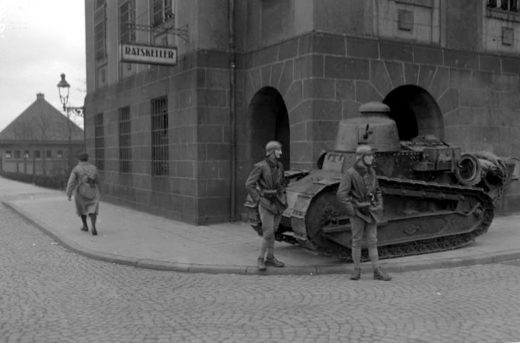 Schwarz-weiß-Bild, ein Eckhaus, vor dem ein Panzer und mehrere Soldaten stehen.